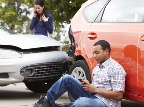 Two cars have been in a fender-bender. A woman stands, talking into a cell phone. A man sits on the ground looking at his cell phone.