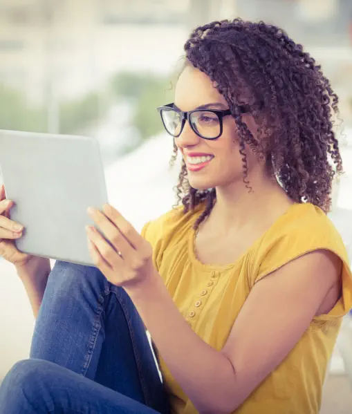A young woman with curly hair and glasses  is smiling and looking at a tablet