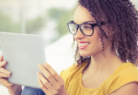 A young woman with curly hair and glasses  is smiling and looking at a tablet